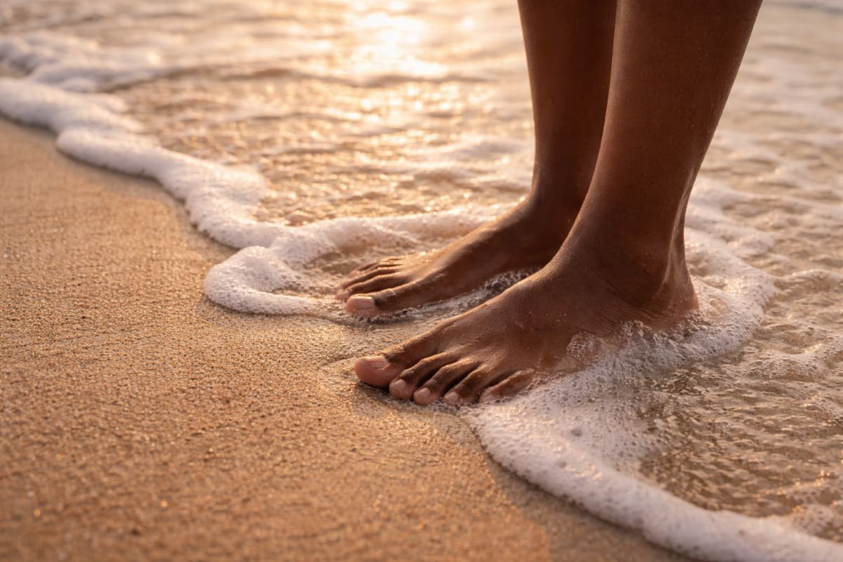 Black woman’s feet in the sand as gentle ocean waves touch the shore